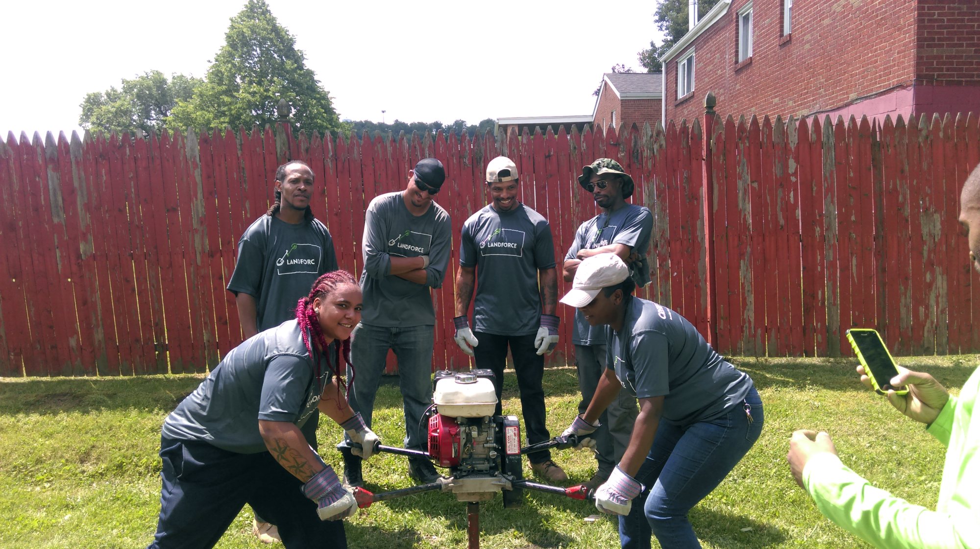 A Landforce crew in grey T-shirts and gloves works a two-person gas auger to dig a planting hole in a fenced backyard lawn while teammates look on