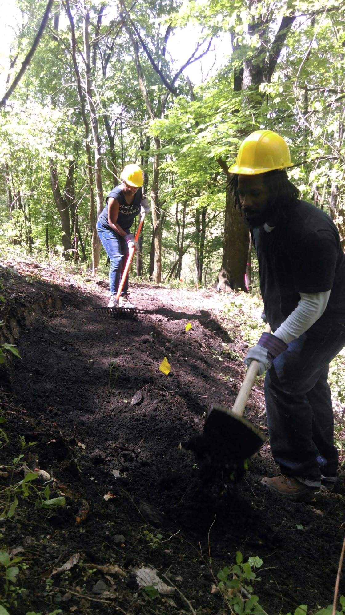 A crew member in a yellow hard hat digs a new trail into a steep wooded hillside, a second crew member working above