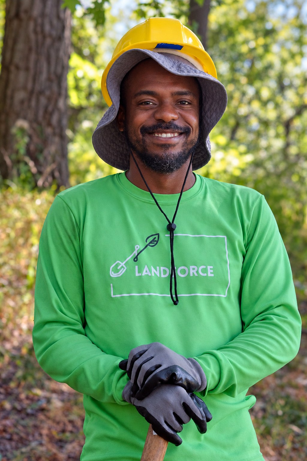A smiling crew member in a green Landforce long-sleeve shirt, yellow hard hat and work gloves on a wooded trail