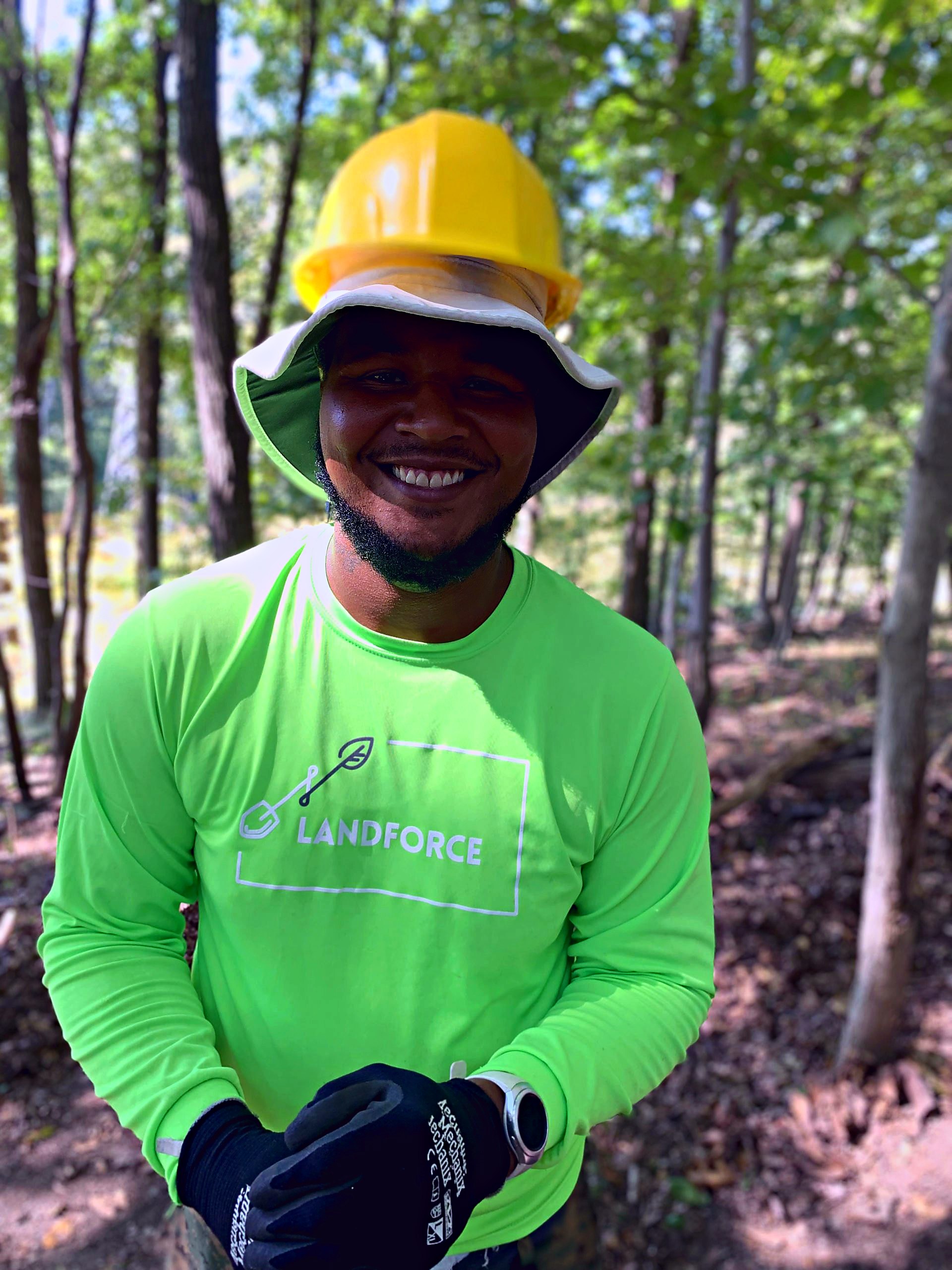 A crew member laughs on a wooded trail, yellow hard hat and green Landforce shirt