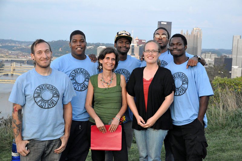 Seven members of the original team, most in matching light-blue trail corps T-shirts, pose on a hillside with the Pittsburgh skyline behind
