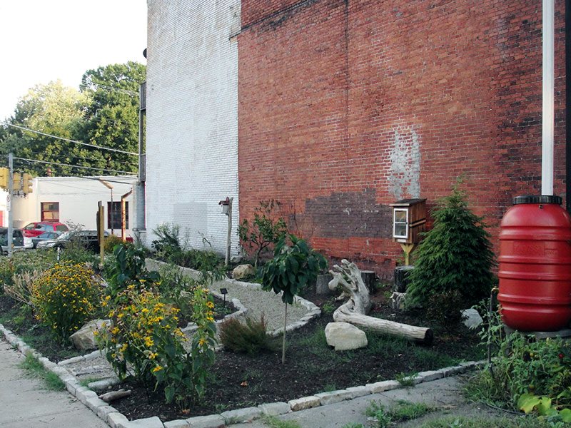 The same lot after work: planted beds, shrubs, edging stones and a red rain barrel along the brick wall