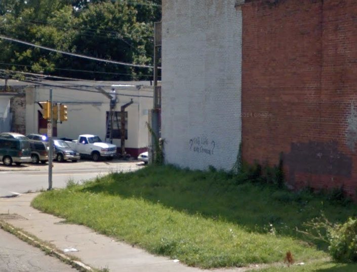 A vacant corner lot before work: patchy grass and a bare sidewalk edge beside a tall brick wall