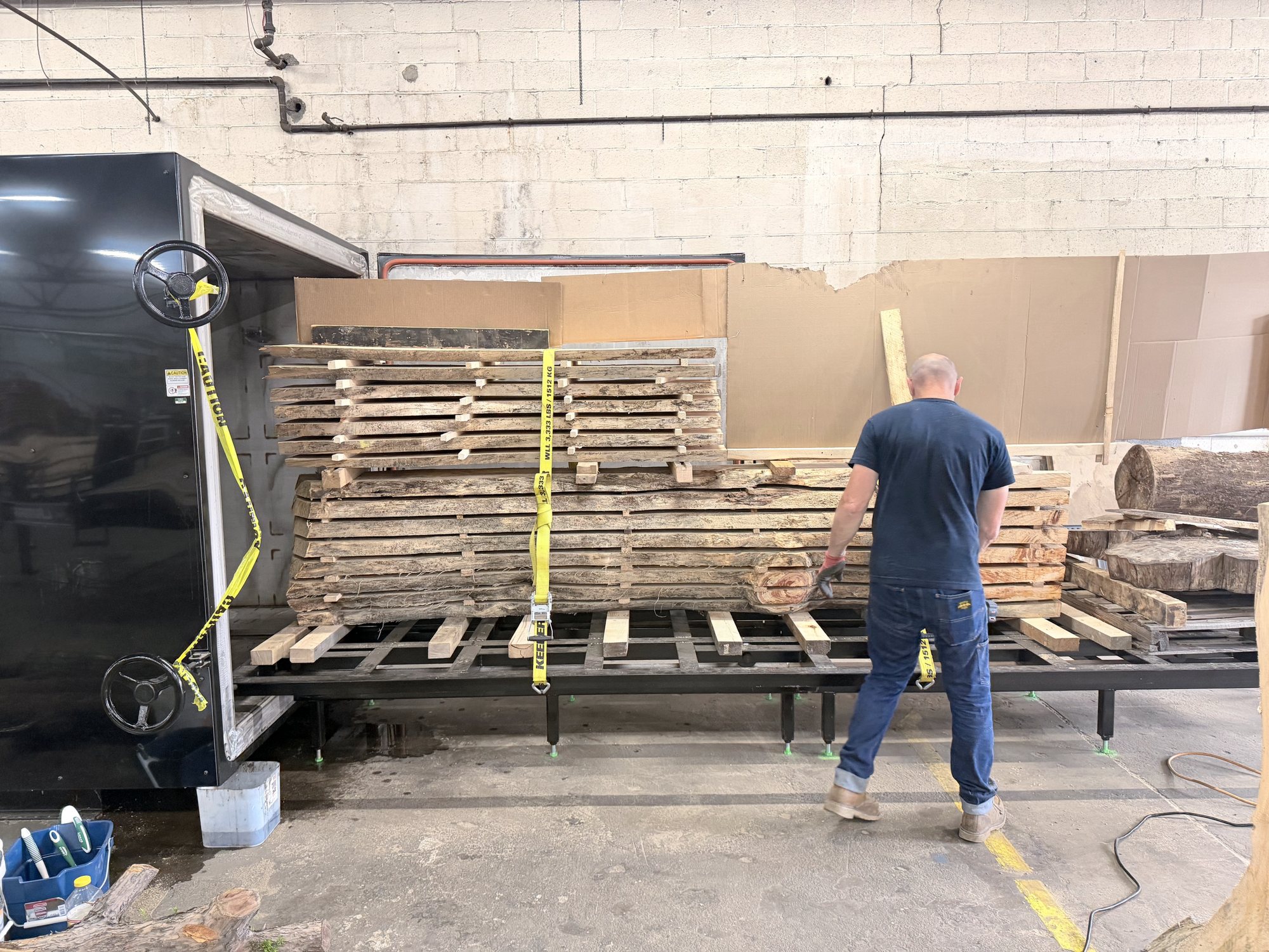 Stacks of lumber strapped onto a rolling cart at the open door of a large black drying chamber, a worker beside it