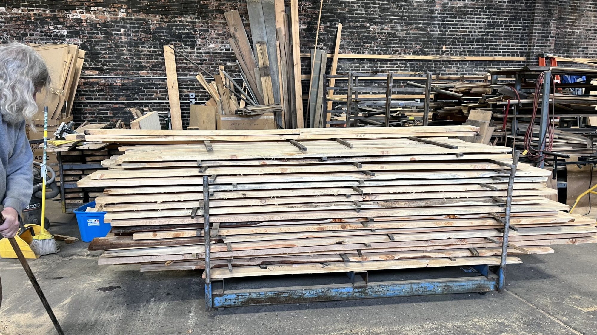 Rough-sawn boards stacked flat on a steel rack in a workshop, more lumber leaning against the brick wall behind