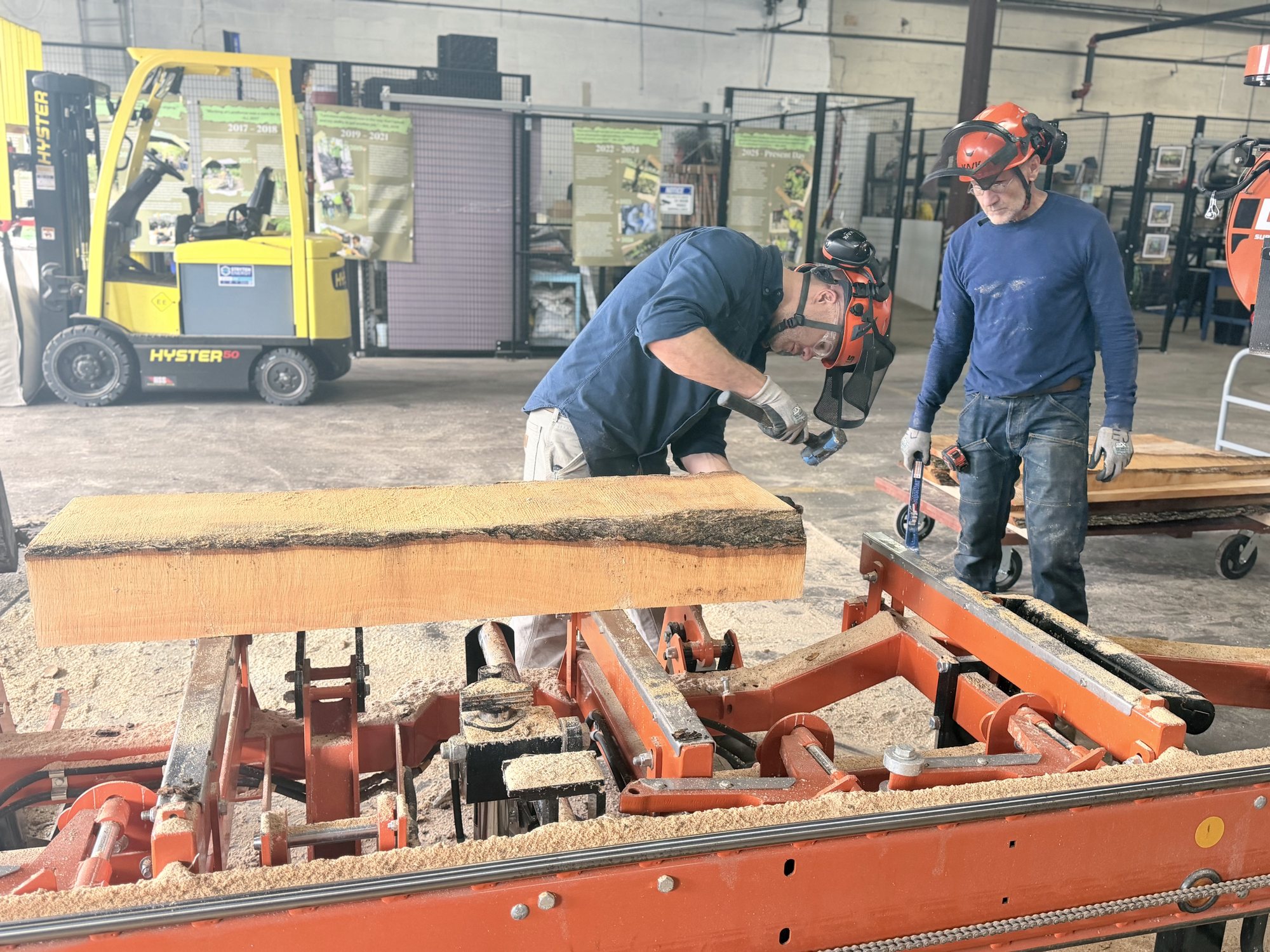 Two workers in hearing protection guide a freshly cut board off the orange Wood-Mizer sawmill, a forklift behind them