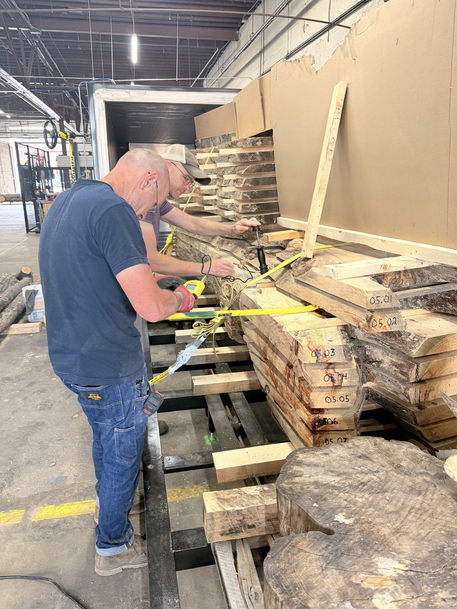 A worker in a blue T-shirt straps a stack of sawn boards on a pallet, a large log round in the foreground