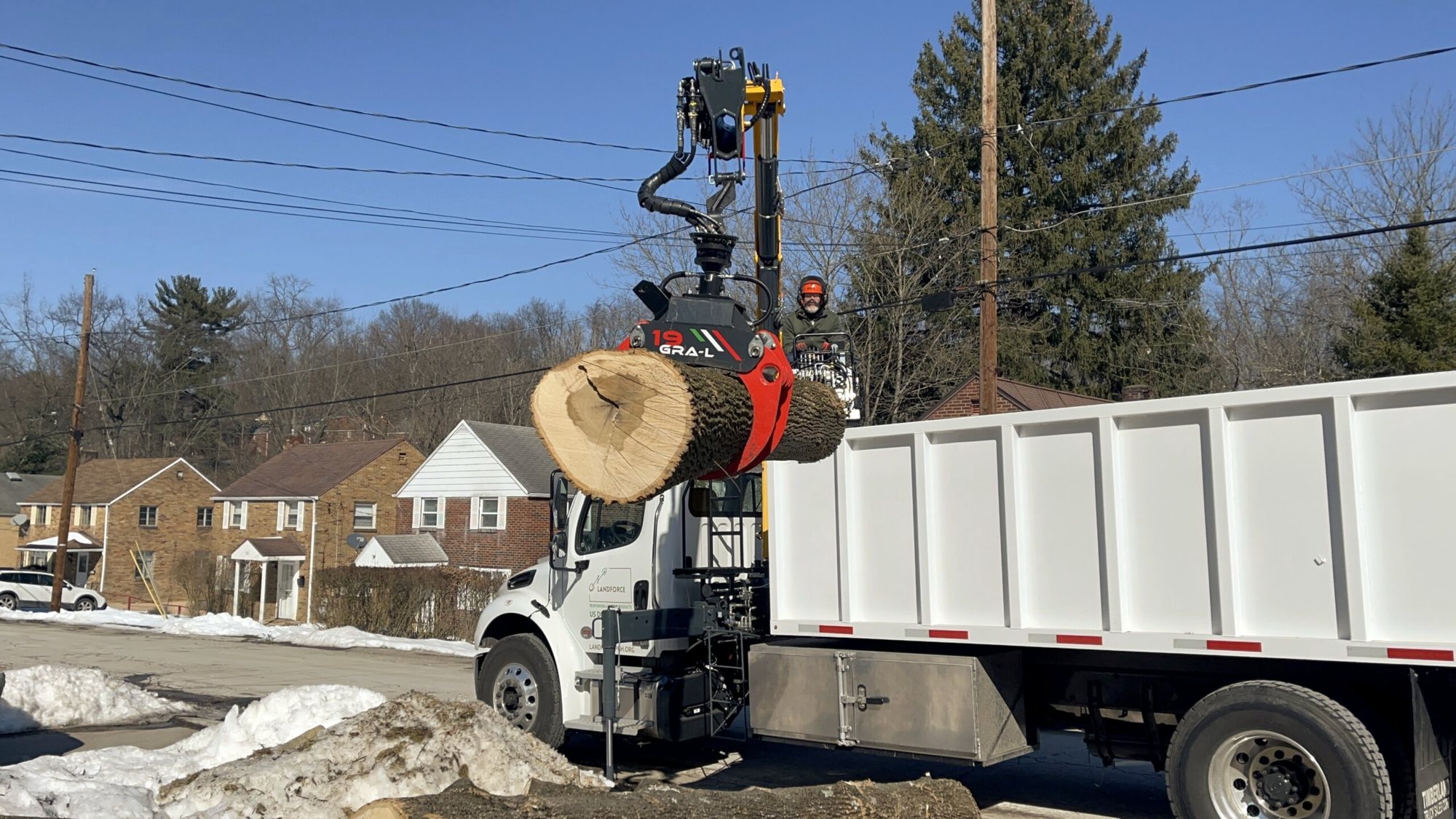 Landforce’s white grapple truck lifts a large log section with its crane beside a residential street, patches of snow on the ground