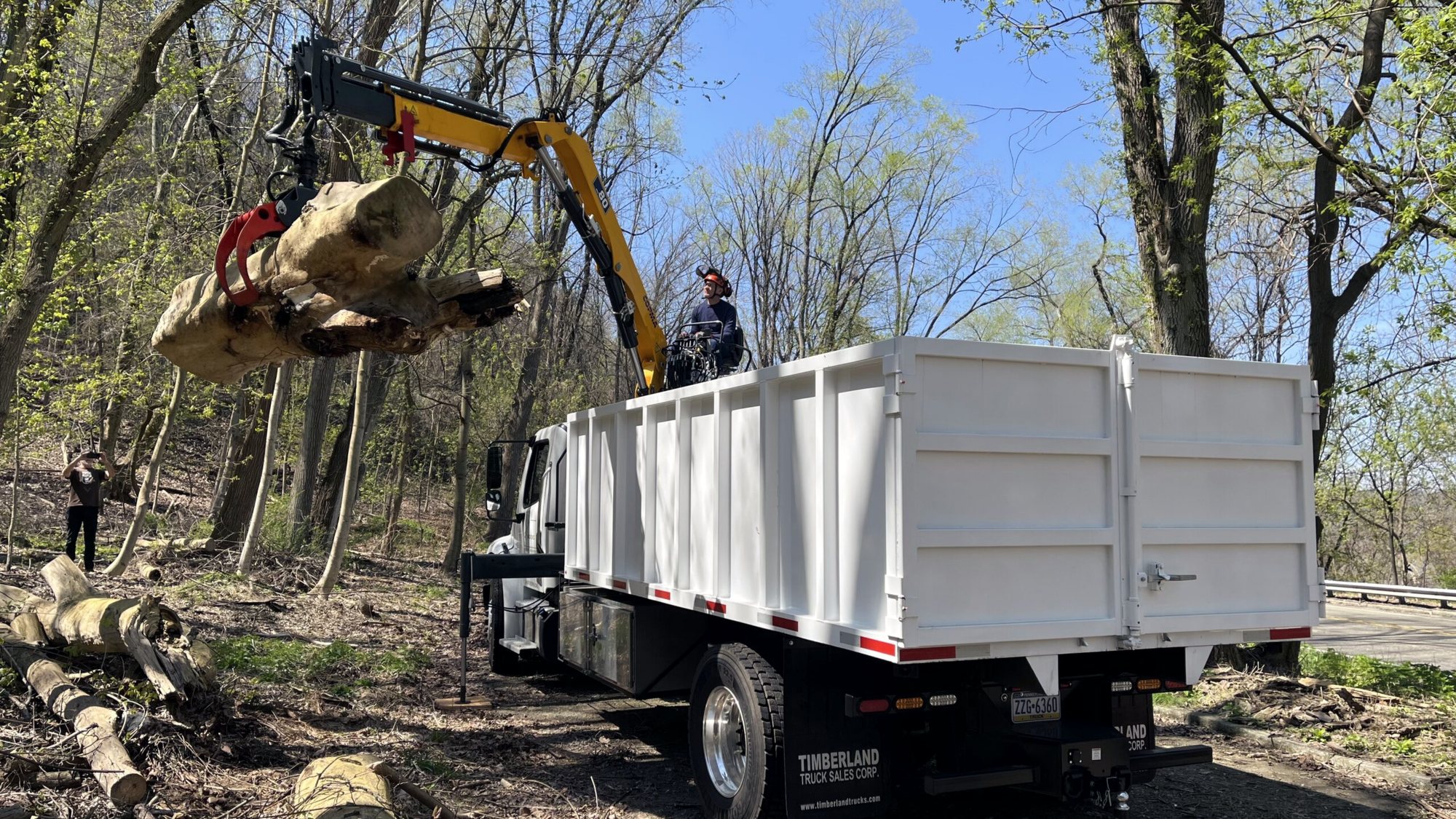 The grapple truck’s crane swings a log into the white dump body at a wooded recovery site