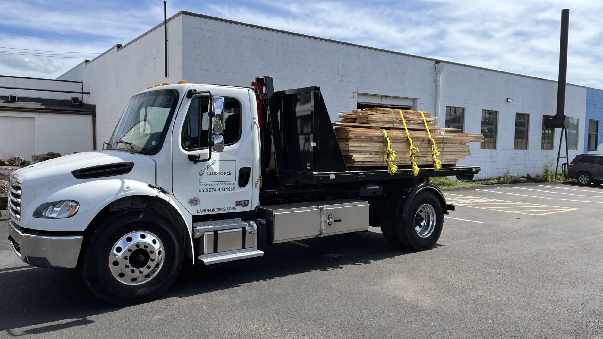 A white flatbed truck carrying strapped-down stacks of sawn lumber, parked beside a large white industrial building
