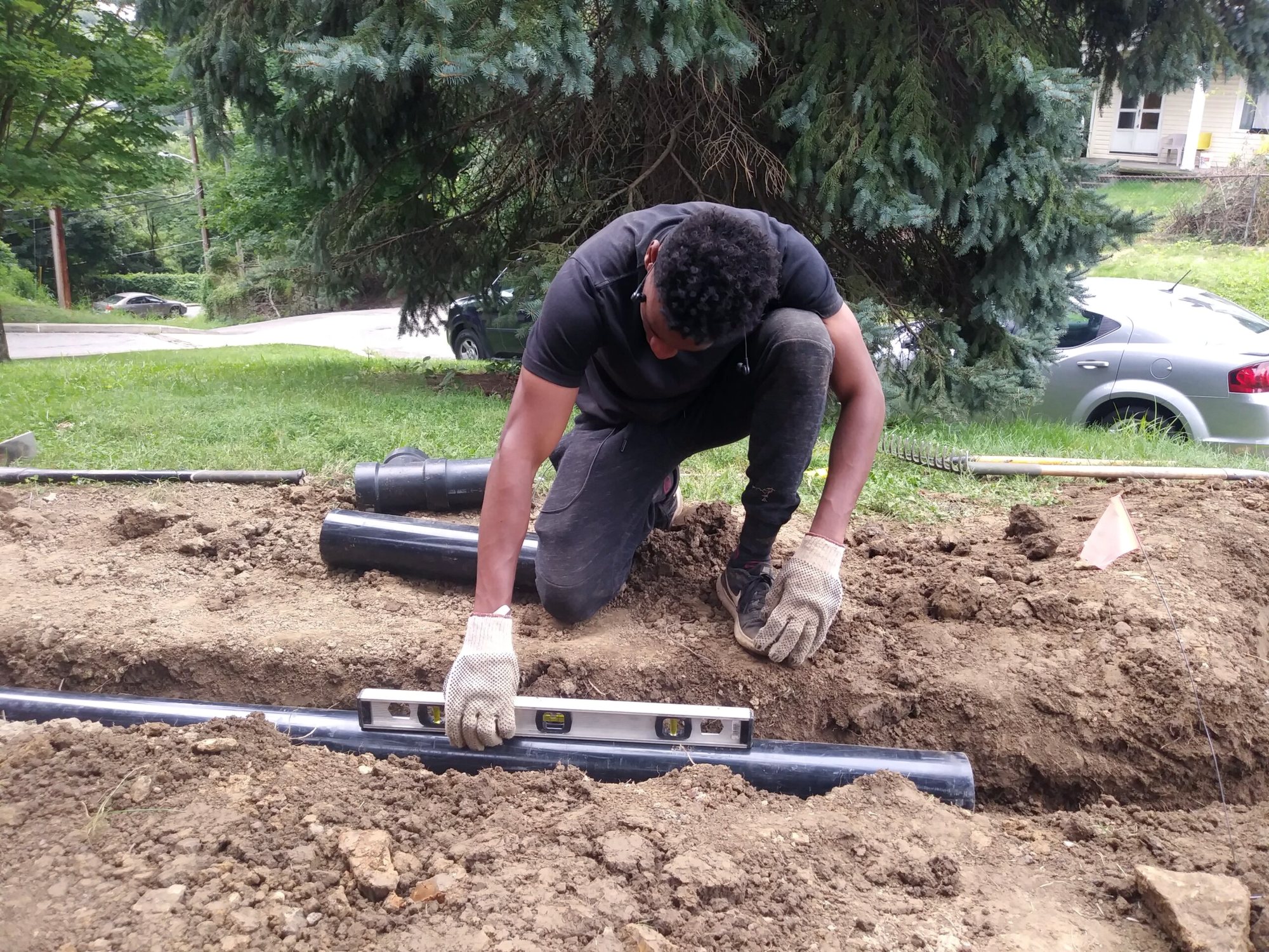A crew member kneels in an excavated trench, checking a black drainage pipe with a spirit level