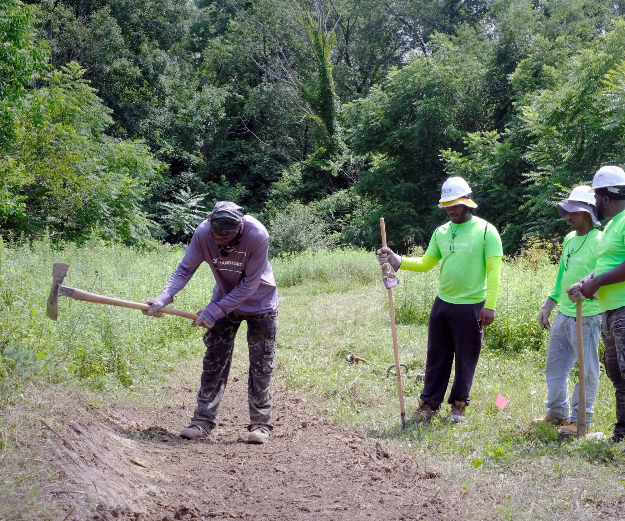 A crew member swings a pick-mattock to break ground in a meadow while crew members in bright green shirts and hard hats look on
