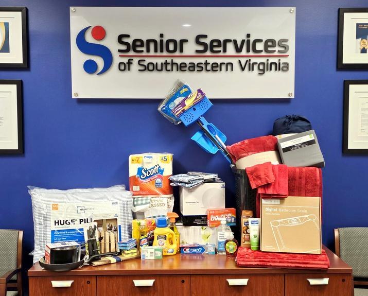 A Welcome Home Basket of bedding, towels, cookware, cleaning supplies and toiletries laid out beneath the Senior Services sign